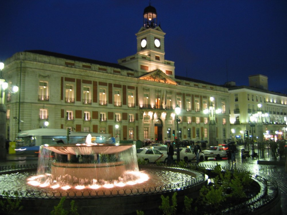 Vista nocturna de la Puerta del Sol de Madrid, con la torre del reloj iluminada del edificio histórico al fondo. En primer plano, una fuente circular iluminada, rodeada de coches y gente.