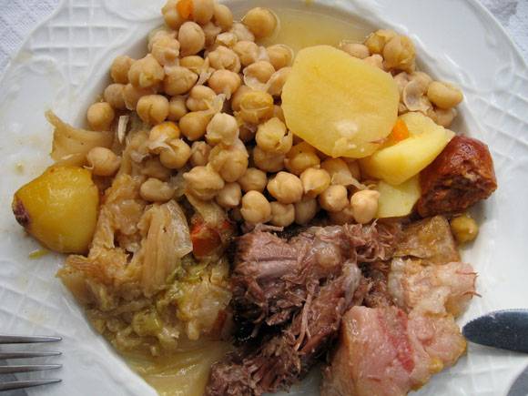 Un plato de estofado con garbanzos, patatas, col y trozos de carne, servido en un plato blanco. La comida parece abundante, con un caldo visible que cubre los ingredientes, lo que sugiere un plato sabroso y reconfortante. Se ve parcialmente un tenedor a un lado.