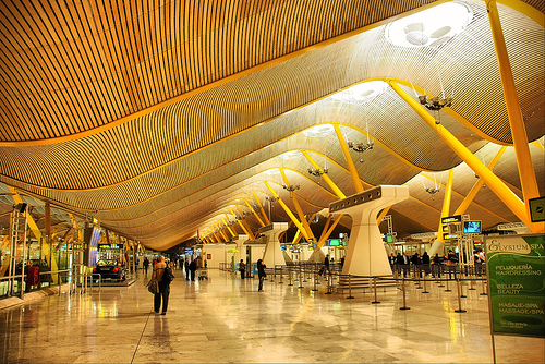 Amplia terminal del aeropuerto con techo ondulado de madera y columnas amarillas. La gente camina y factura en los mostradores. La brillante iluminación realza el moderno diseño arquitectónico. La señalización indica las zonas de servicio y se ve una fila de pasajeros esperando.