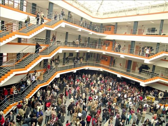 Un gran atrio con público en varios niveles. El espacio cuenta con escaleras y barandillas naranjas y grises. La planta principal acoge a una multitud densa, mientras que otros se encuentran en los balcones de los niveles superiores. El techo es de cristal.