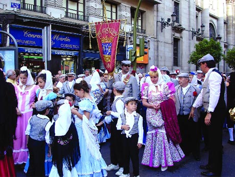 Una multitud de personas con atuendos tradicionales y coloridos se reúne en una calle de la ciudad durante un desfile. Adultos y niños, algunos con sombreros y vestidos florales, se encuentran frente a tiendas con pancartas. El ambiente es festivo y animado.